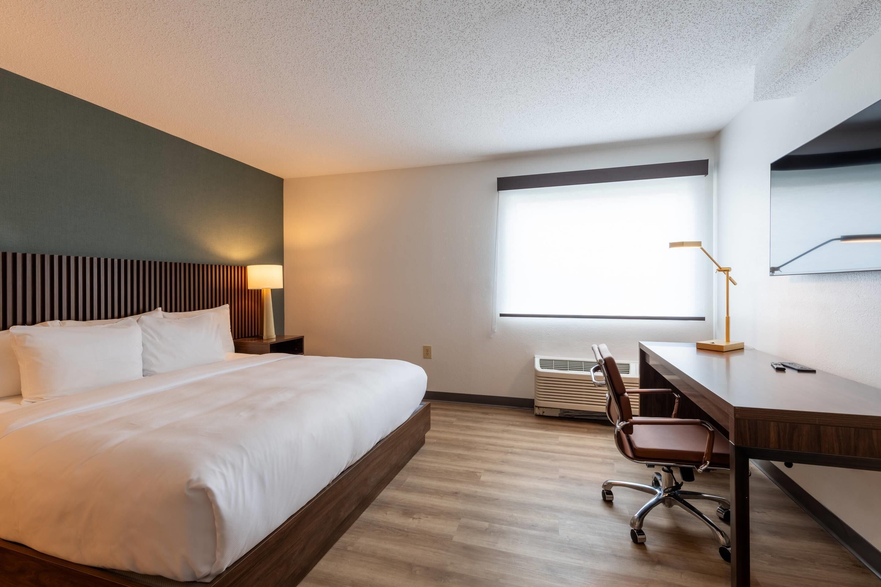 Habitación de hotel con cama, escritorio, silla y un ventanal amplio.