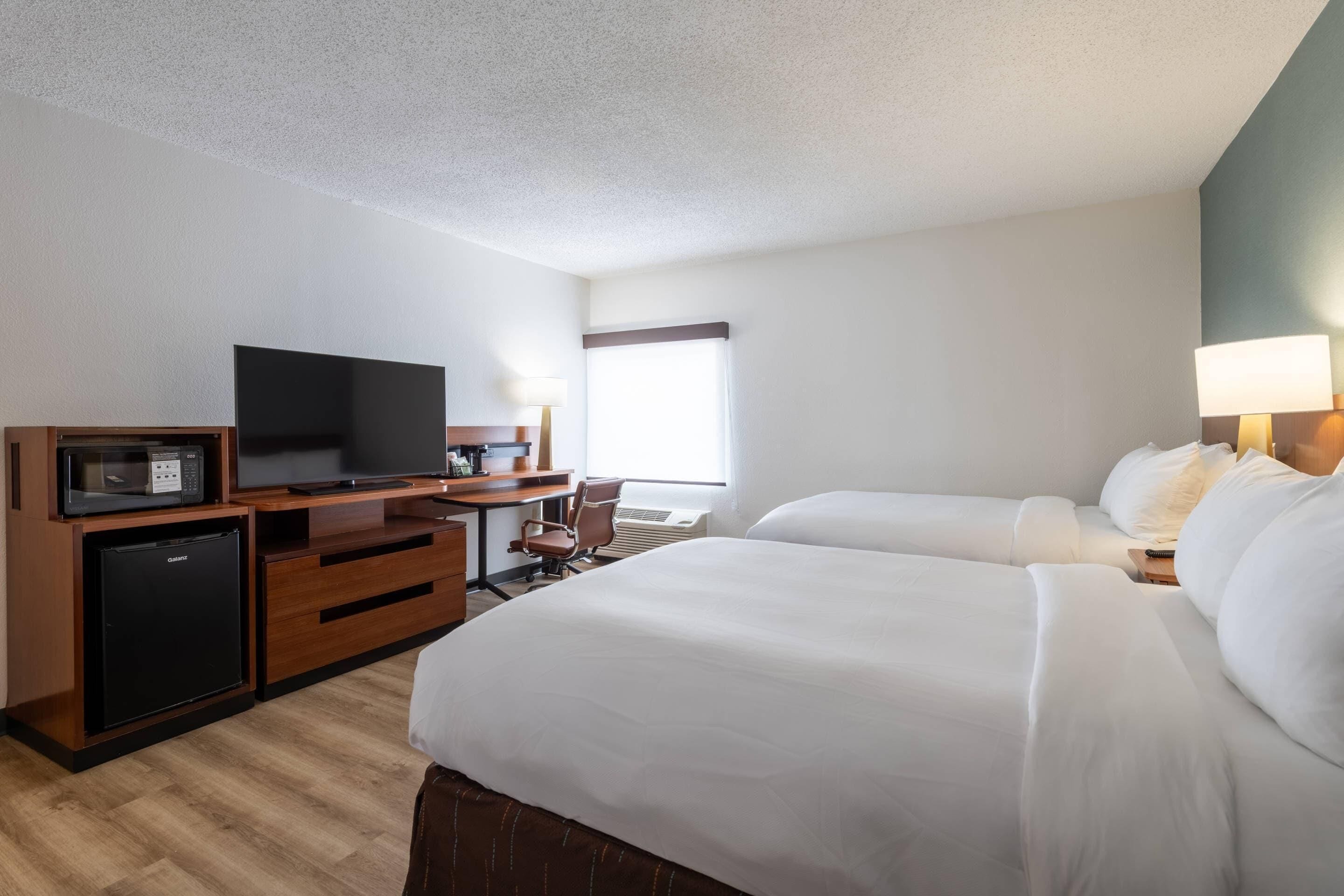 Ein Hotelzimmer mit Flachbildfernseher, kleinem Kühlschrank, Schreibtisch mit Stuhl und zwei Betten mit weißer Bettwäsche.