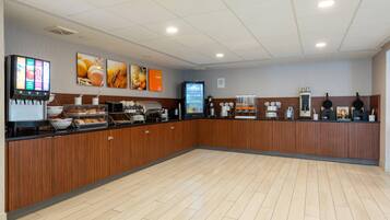 Un espace pour le petit-déjeuner avec un buffet où les clients peuvent se servir eux-mêmes, des machines à café et un écran numérique affichant le menu.