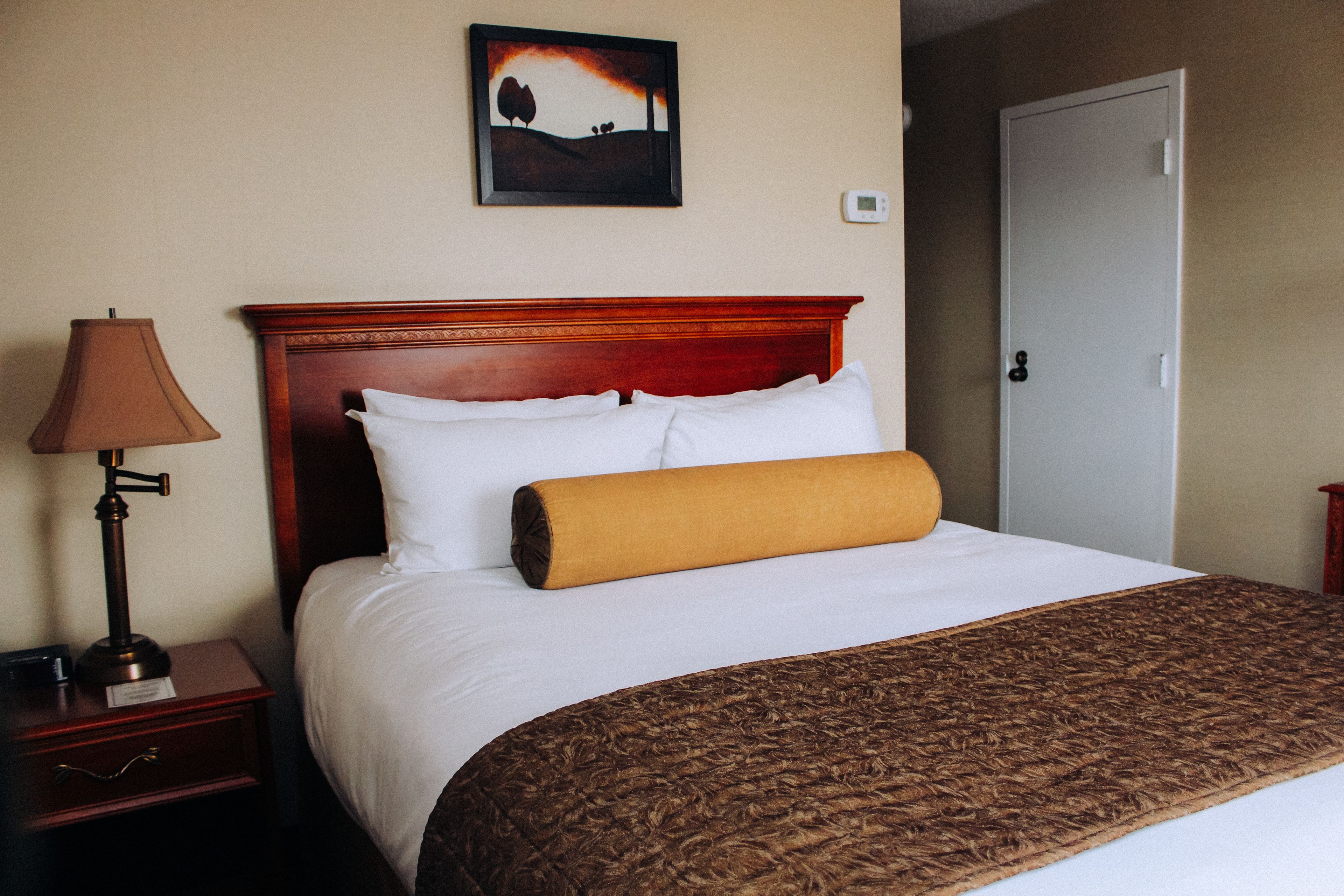 Habitación de hotel con cabecera de madera, ropa de cama blanca, manta marrón, una lámpara y un cuadro en la pared.