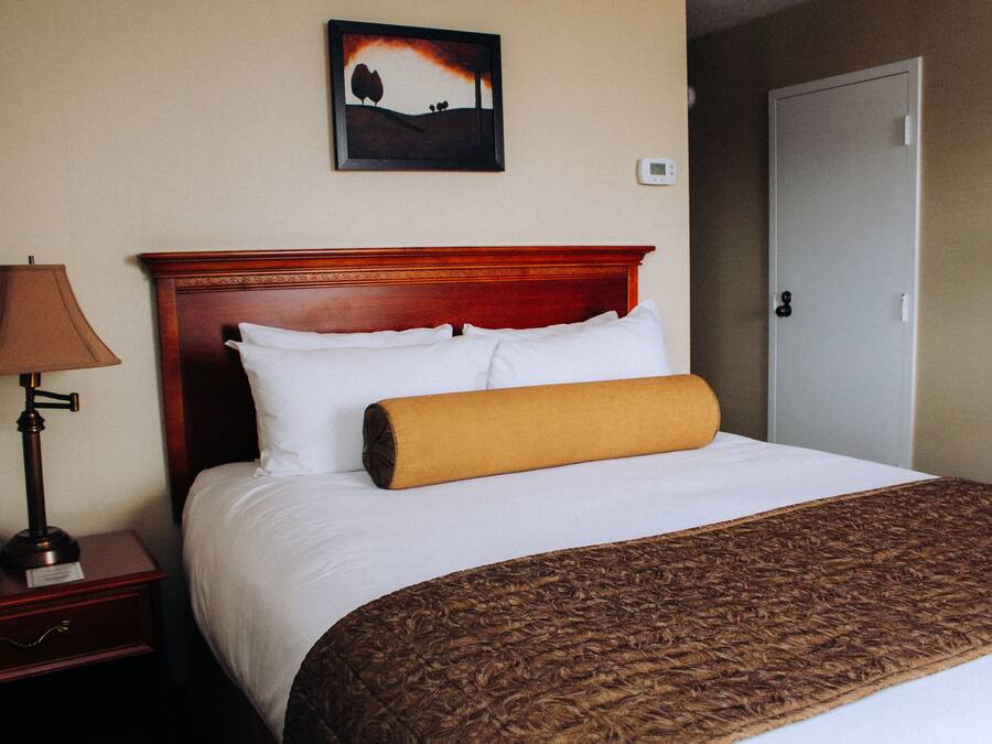 A hotel room with a wooden headboard, white bedding, a brown throw blanket, a lamp, and a painting on the wall.
