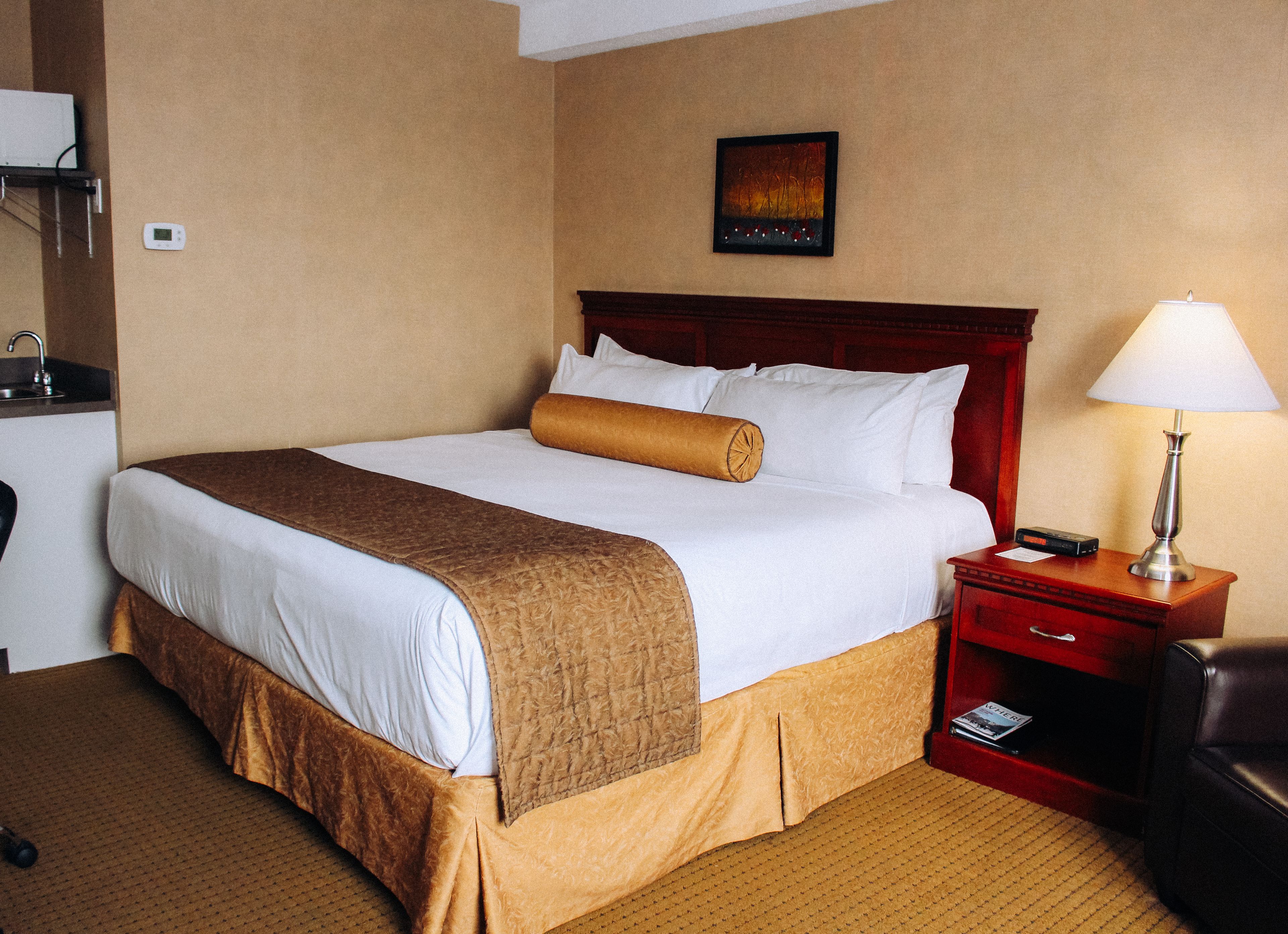 A hotel room with a large bed, a bedside table with a lamp, a small desk with a telephone, and a wall-mounted thermostat.