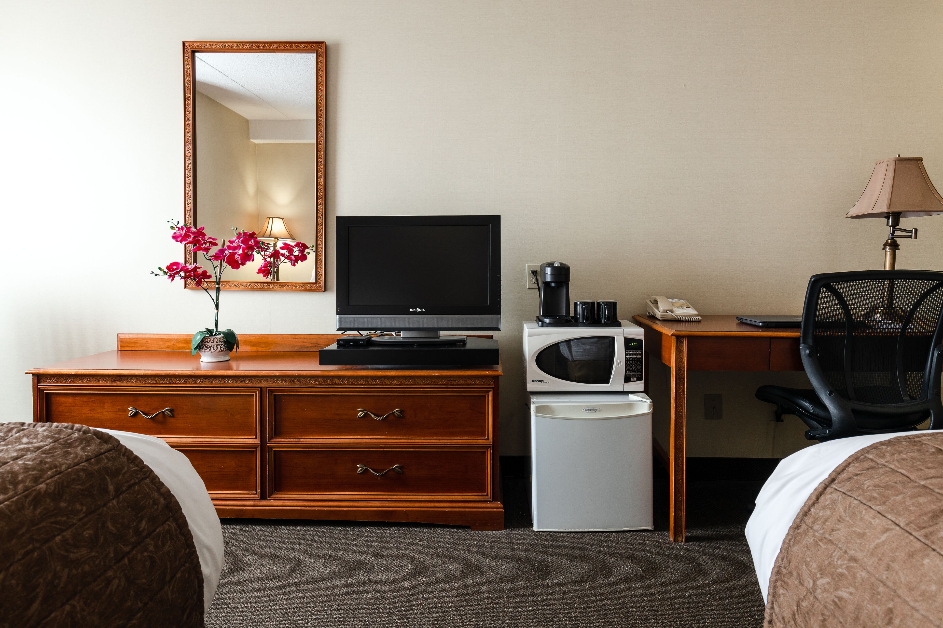 A hotel room with a wooden dresser, a television, a microwave, a desk with a lamp, and two beds.