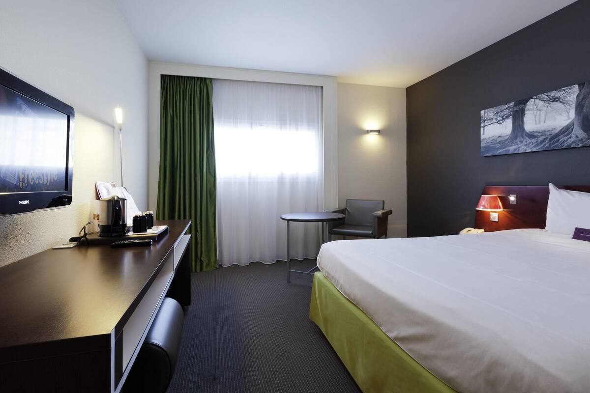 A hotel room with a bed, a desk with a coffee maker, a chair, a small table, a TV, and a painting on the wall.