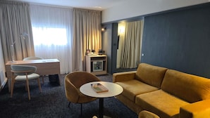 Ein Hotelzimmer mit einer Couch, zwei Sesseln, einem kleinen runden Tisch, einem Schreibtisch und einer Lampe.