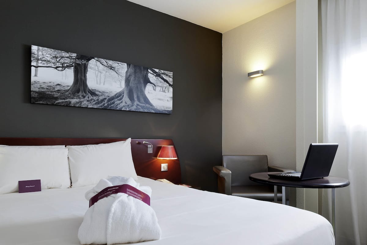 A hotel room with a bed, a desk with a laptop, a wall-mounted lamp, and a painting of trees.