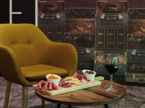 Un sillón color mostaza, una mesa de madera con una tabla de quesos, una copa de vino tinto y maletas antiguas al fondo.
