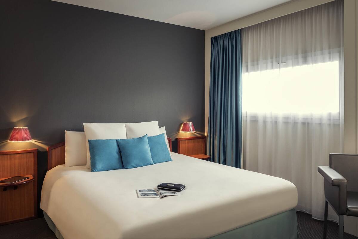 A hotel room with a large bed, two bedside tables with lamps, a chair, and a window with curtains.