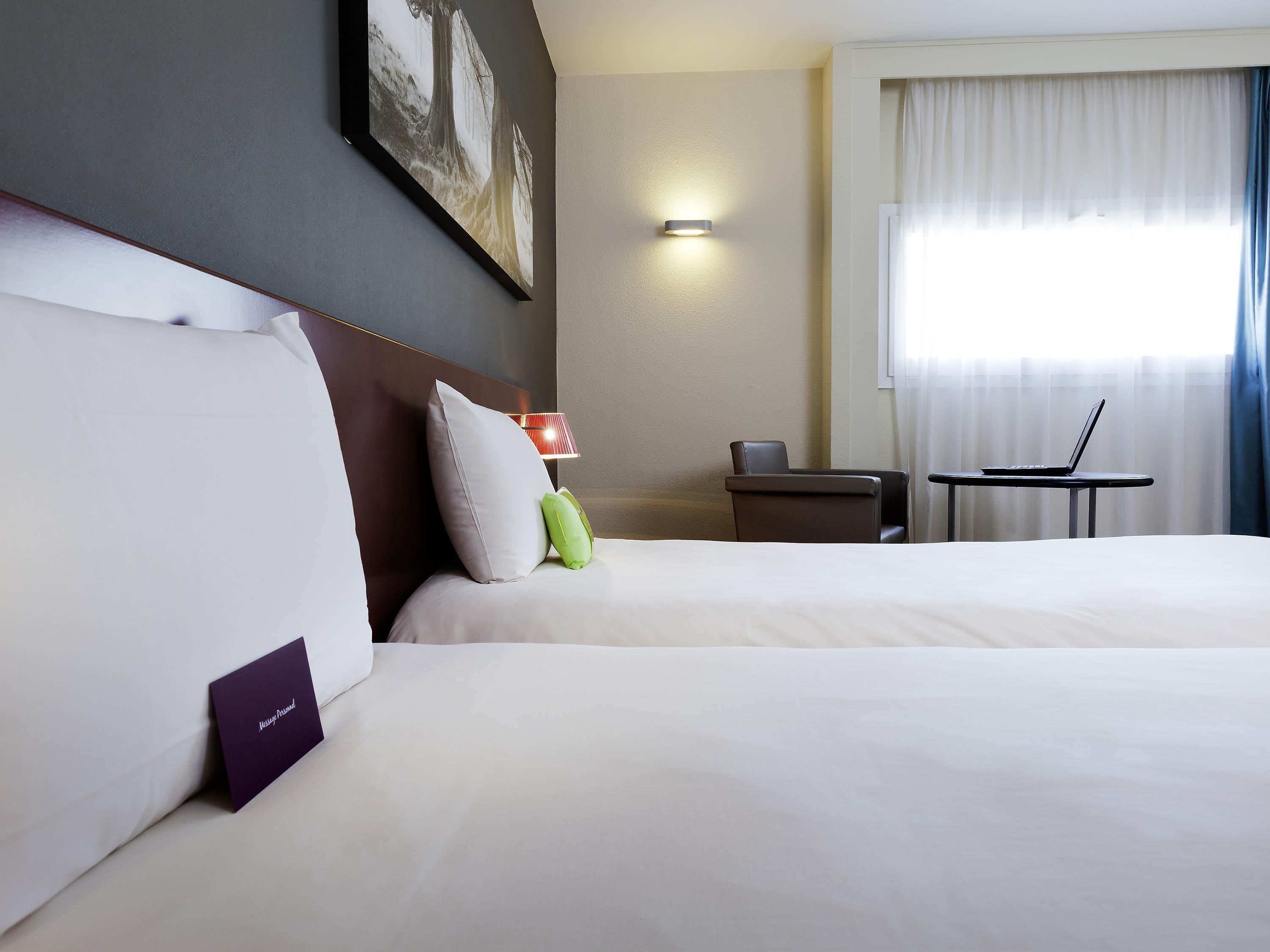 A hotel room with two beds, a desk, a chair, a lamp, and a painting on the wall.