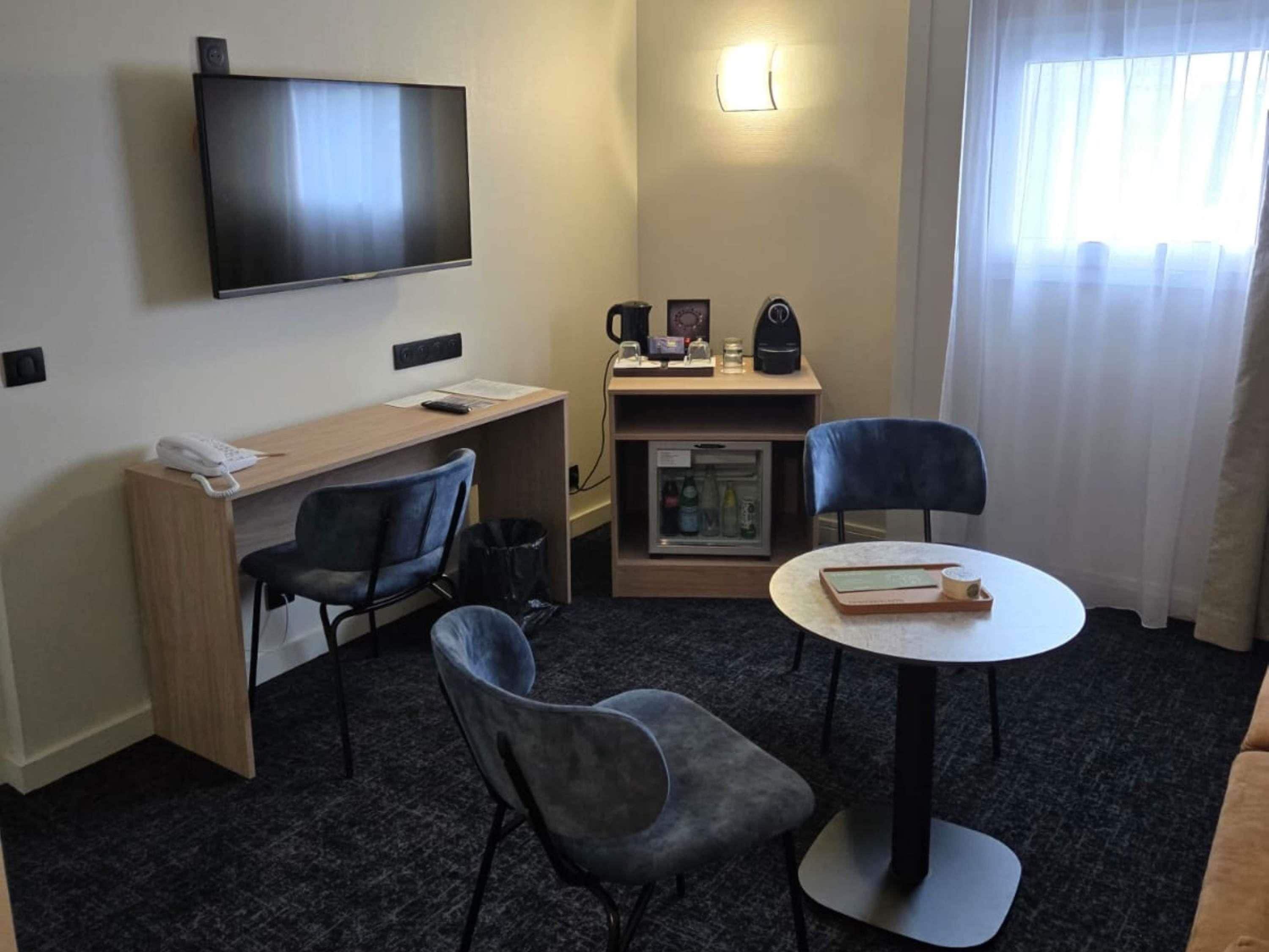 Habitación de hotel con televisor de pantalla plana, escritorio con teléfono, dos sillas, refrigerador pequeño y mesa con un libro y una taza.