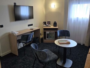 Hotelkamer met flatscreen televisie, bureau met telefoon, twee stoelen, kleine koelkast en tafel met boek en beker.