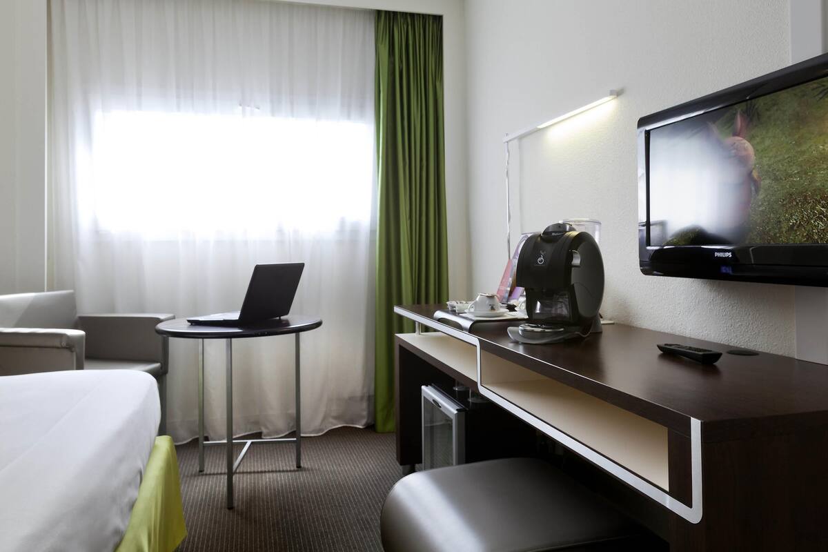 A hotel room with a bed, a desk with a laptop, a coffee maker, a TV, and a green curtain.