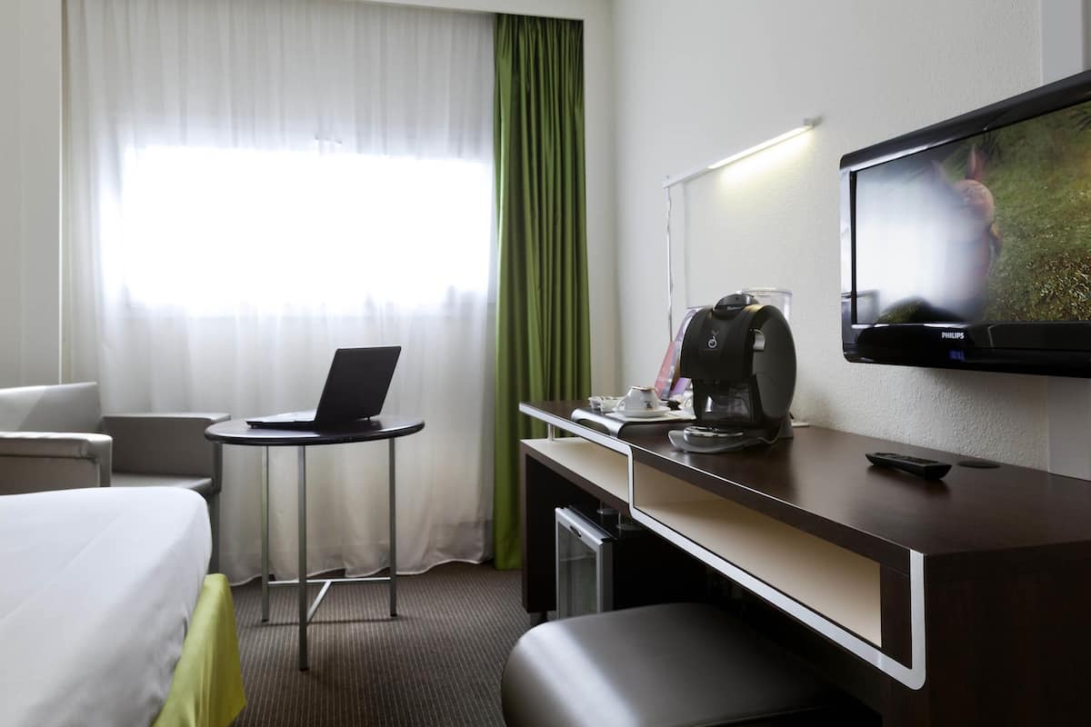 A hotel room with a bed, a desk with a laptop, a coffee maker, a TV, and a green curtain.