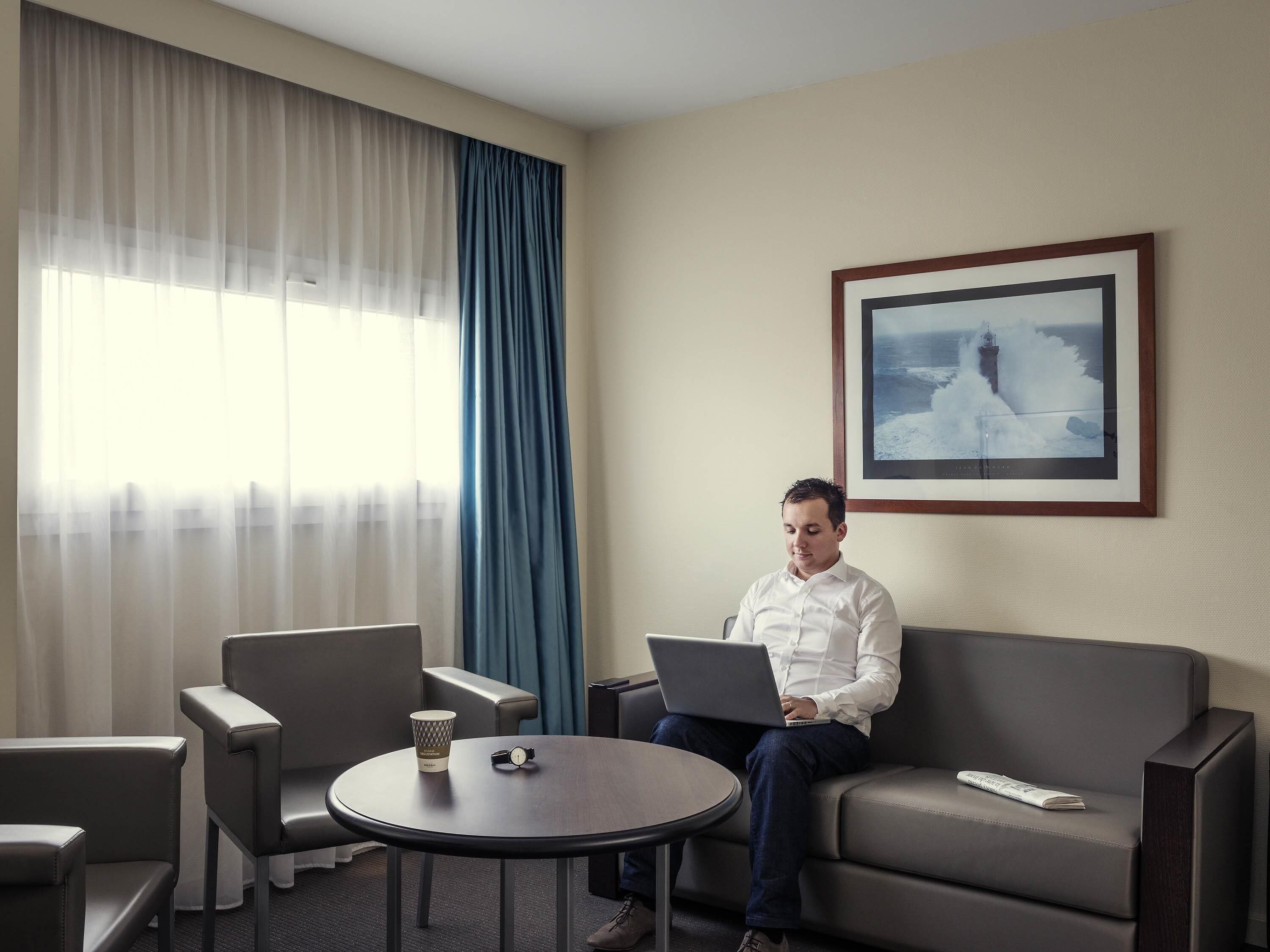 Un homme est assis sur un canapé gris, avec un ordinateur portable, une tasse de café et une montre sur une petite table ronde. On peut voir deux fauteuils gris, une grande fenêtre avec des rideaux légers et un tableau encadré au mur.