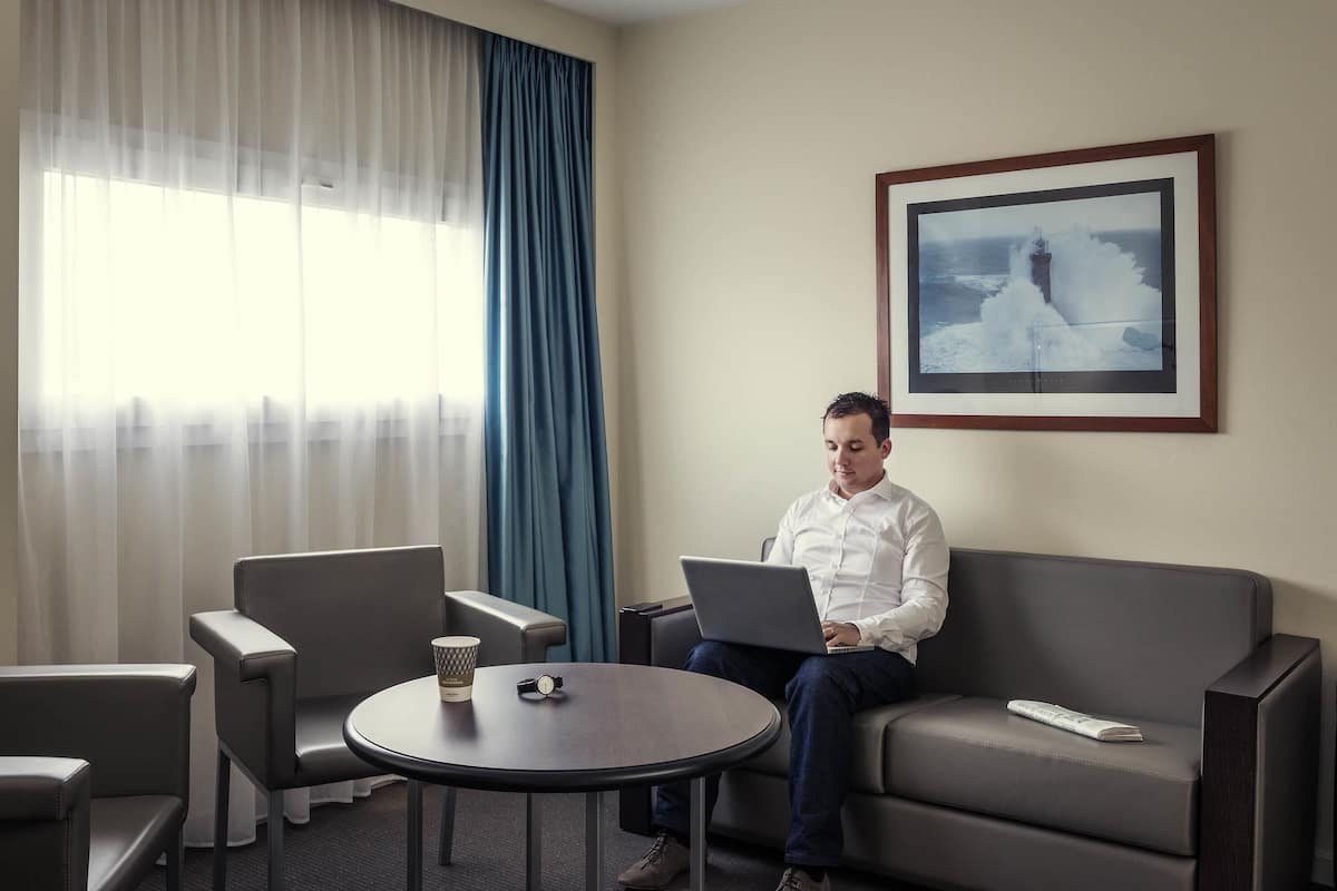 A man sitting on a grey sofa with a laptop, a coffee cup, and a watch on a small round table. There are two grey chairs, a large window with sheer curtains, and a framed picture on the wall.