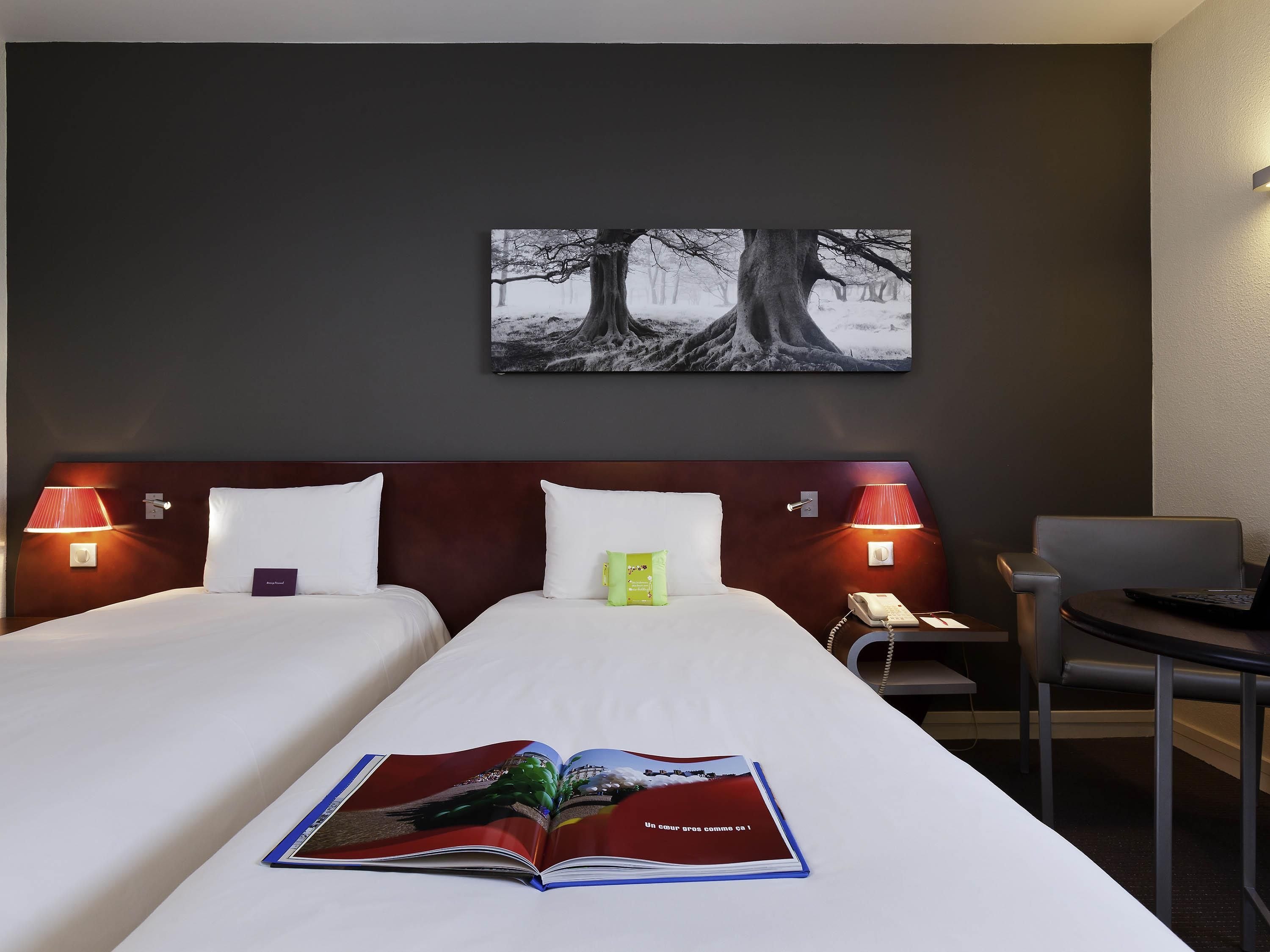 A hotel room with a bed, two bedside lamps, a desk with a laptop, and a painting on the wall.
