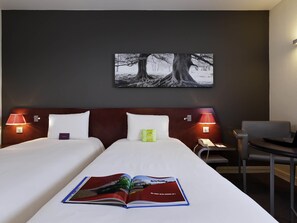 A hotel room with a bed, two bedside lamps, a desk with a laptop, and a painting on the wall.
