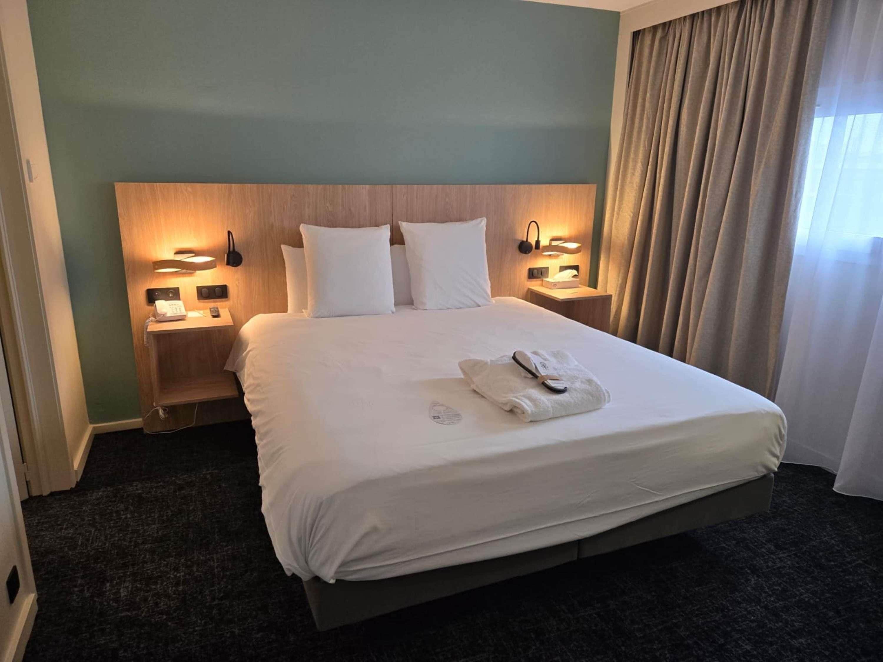 A hotel room with a large bed, two bedside tables, a headboard, and a window with curtains.