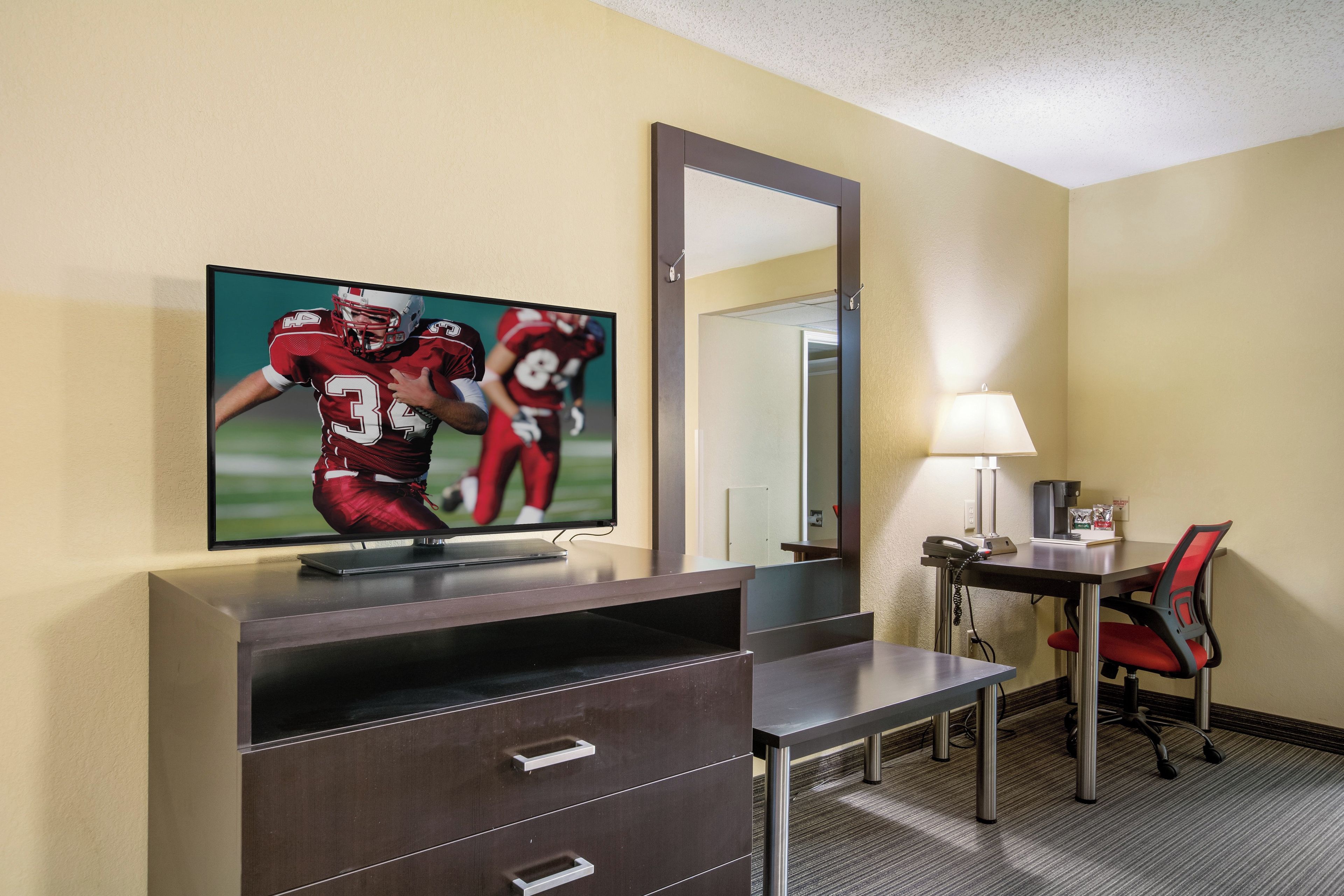 Ein Hotelzimmer mit Fernseher, der ein Sportspiel zeigt, einem Schreibtisch mit Lampe, einem Sessel und einem Spiegel.