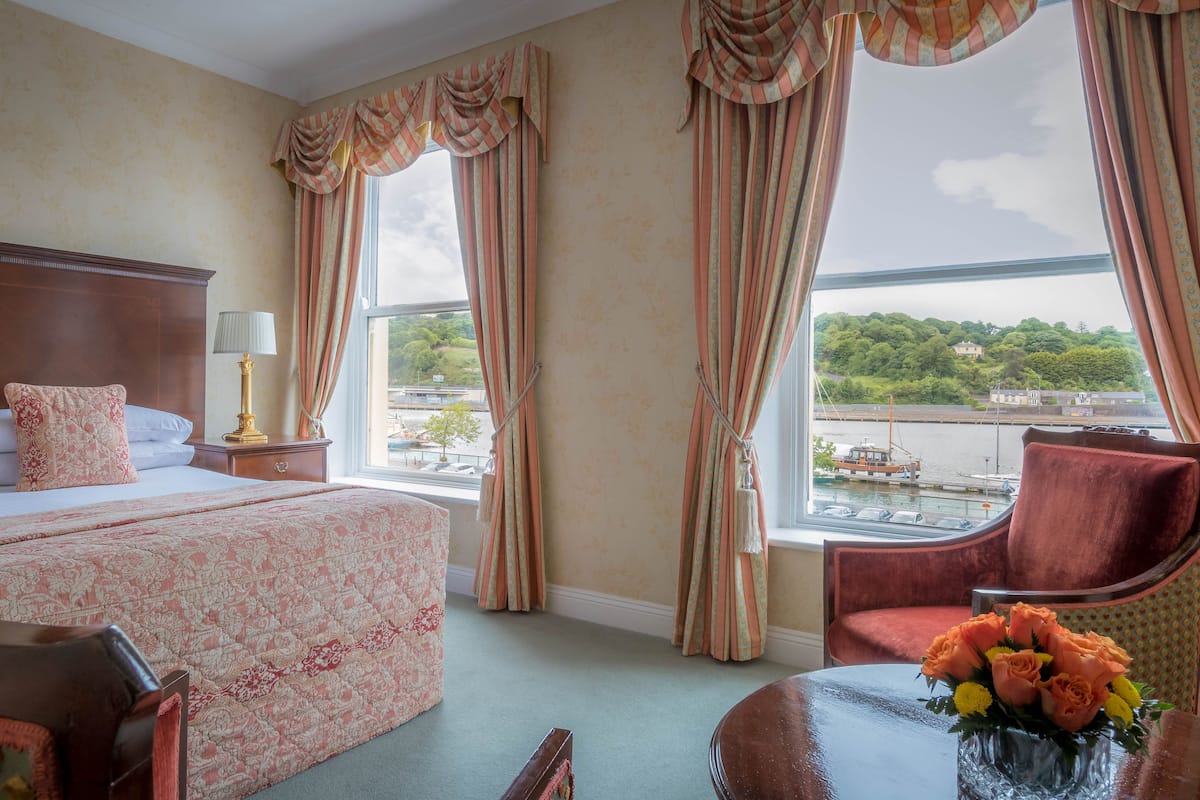 Une chambre d’hôtel avec un grand lit, une table de chevet avec une lampe, une chaise et une vue sur une rivière et la verdure environnante.