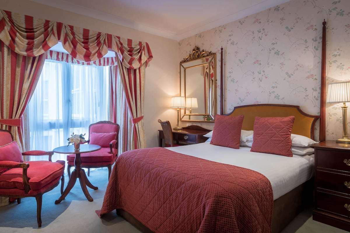 Une chambre d’hôtel avec un grand lit, deux fauteuils rouges, une petite table, une commode et un miroir.