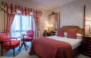 Habitación de hotel con una cama grande, dos sillas rojas, una mesita, un aparador y un espejo.