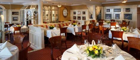 A formal dining area with multiple round tables set for service, featuring white tablecloths, neatly arranged cutlery, and floral centerpieces.