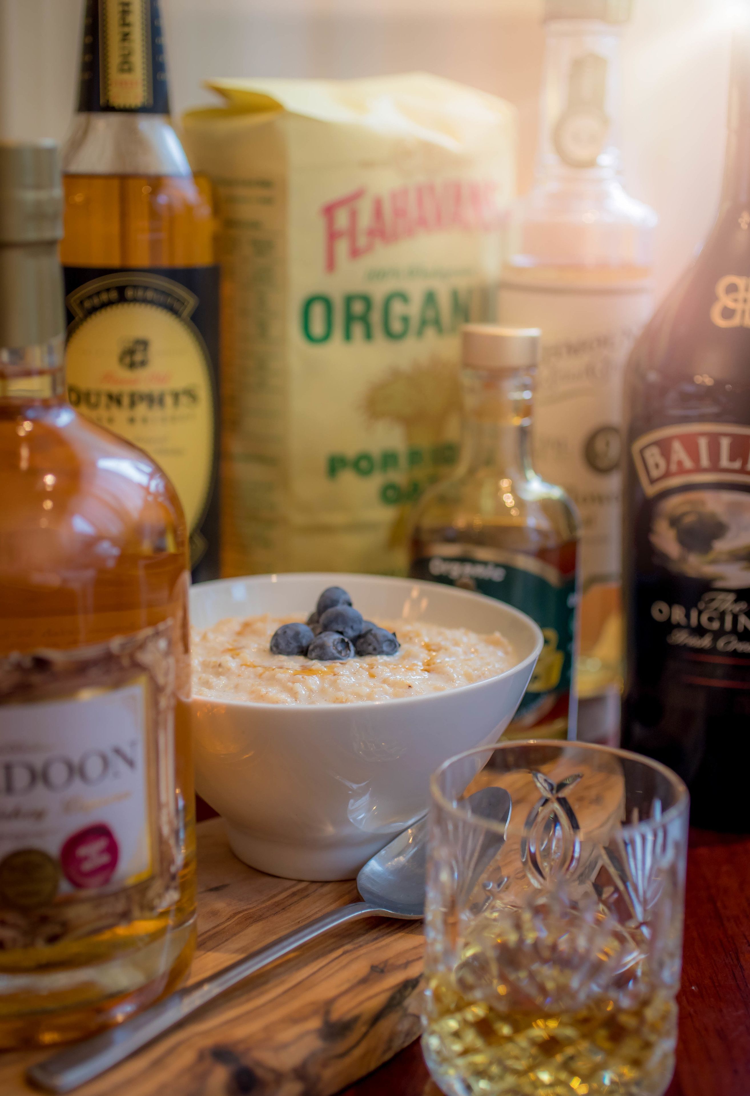 A bowl of oatmeal with blueberries, a glass of whiskey, and bottles of liquor on a wooden table.