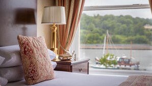 A hotel room with a bed, a nightstand, a lamp, and a view of a lake with boats.