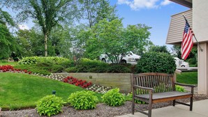 A bench in a landscaped area with a flag, trees, and a building in the background.