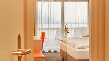 Habitación de hotel con cama, escritorio, sillón naranja, florero con flores y vista a través de cortinas traslúcidas.