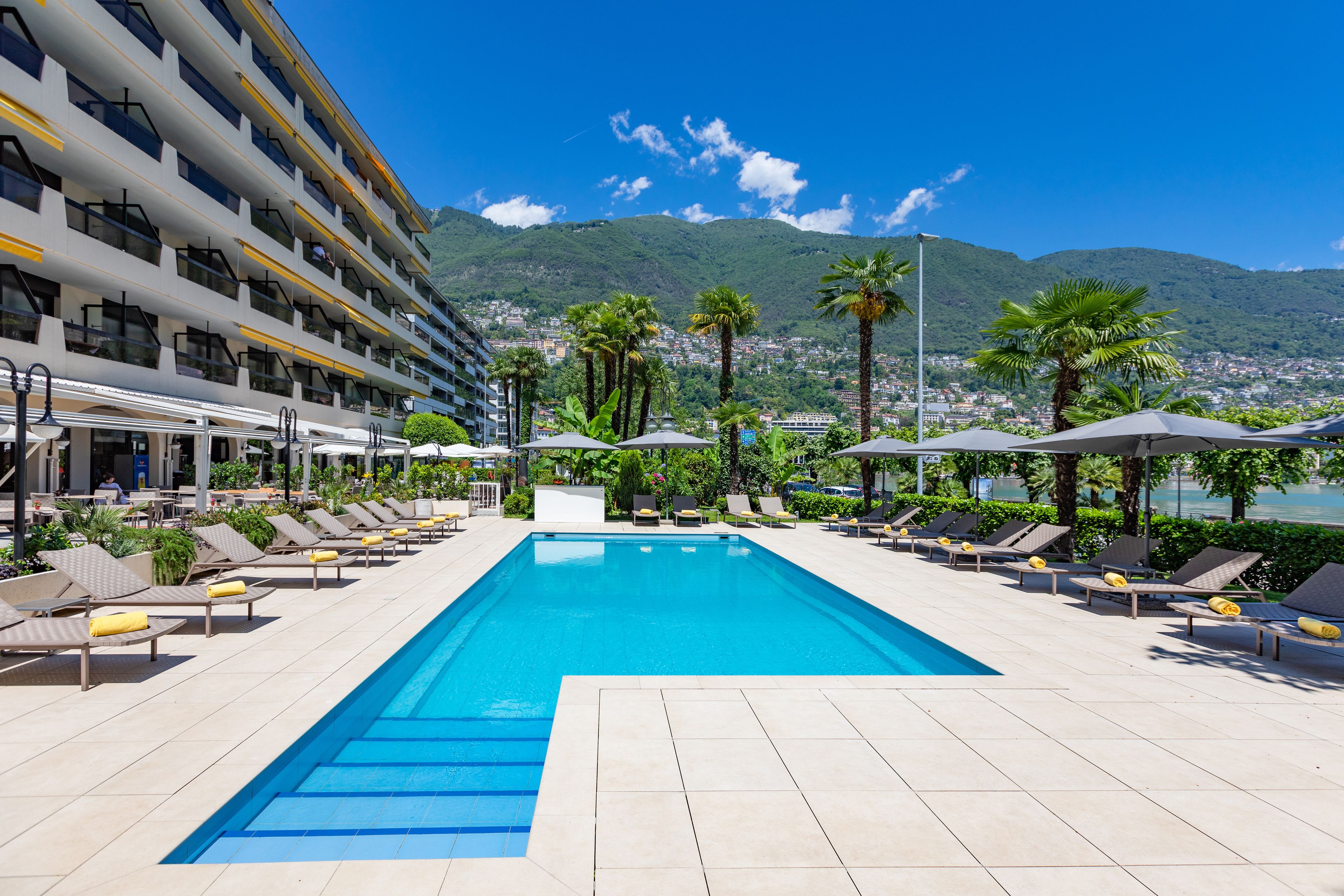 Une piscine dotée de chaises longues et de parasols, entourée d’un bâtiment d’hôtel et offrant une vue sur la montagne.