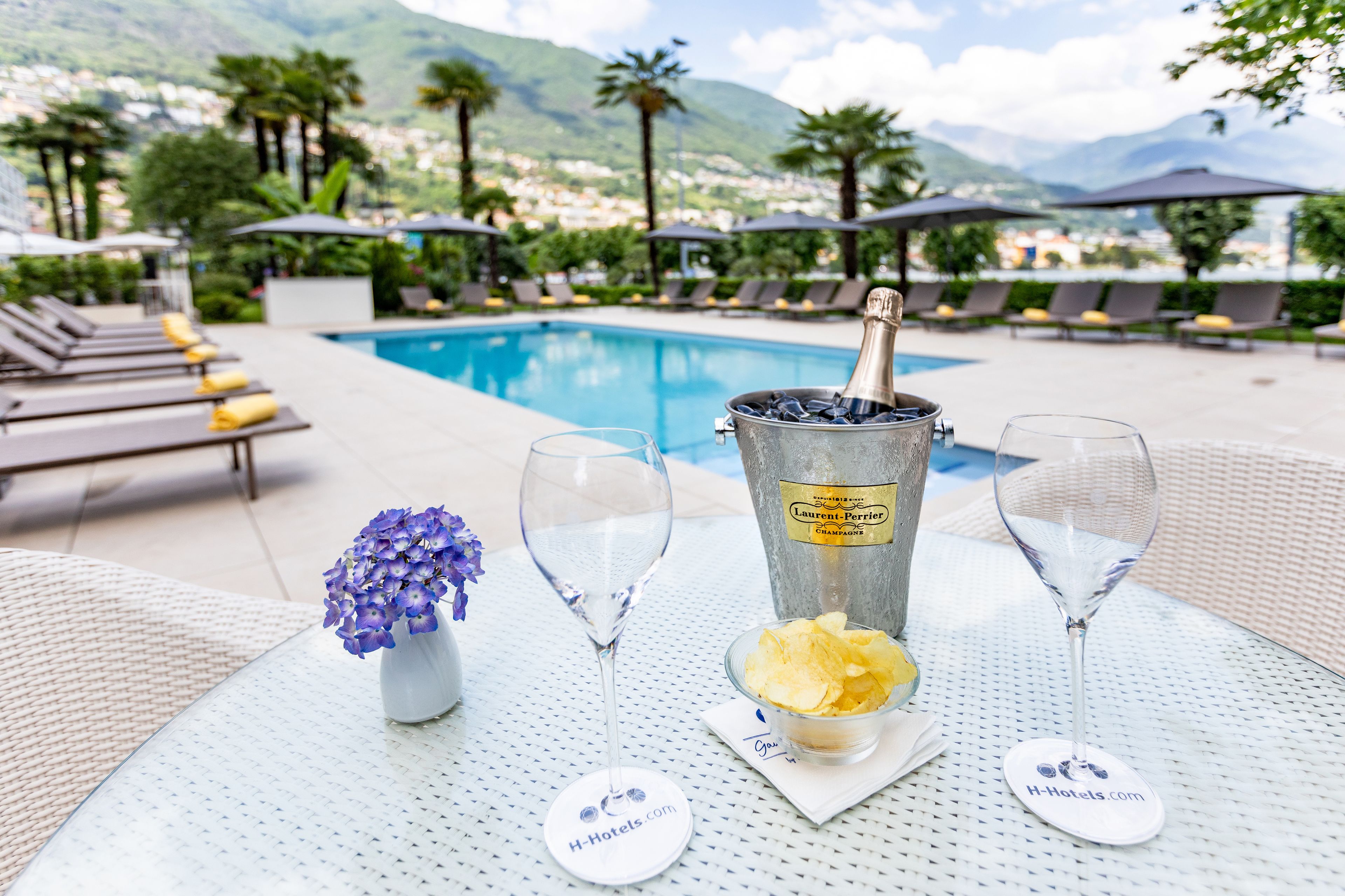 Una mesa junto a la piscina con una botella de champán, dos copas, un florero con flores y un bol de patatas fritas.