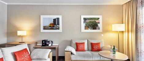 A hotel room with a sofa, a small table, a coffee machine, and two framed pictures on the wall.