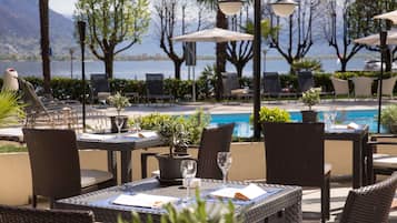 An outdoor seating area with wicker tables and chairs, a small potted plant, and a view of a lake and mountains in the background.
