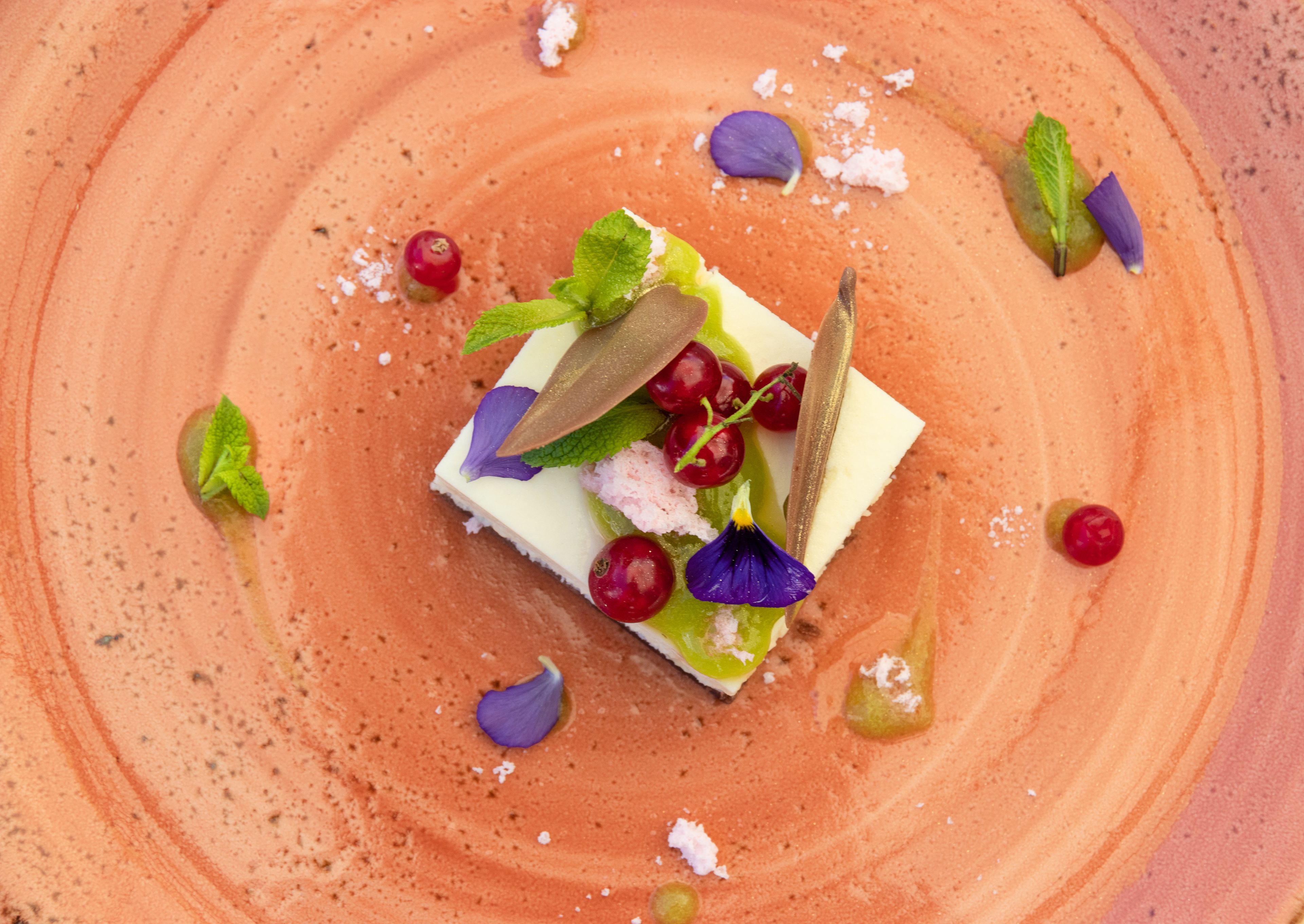 Ein Dessert mit quadratischer Basis, garniert mit Minzblättern, Beeren und einem dekorativen Element auf einem tellerartigen, strukturierten orangefarbenen Untersetzer.
