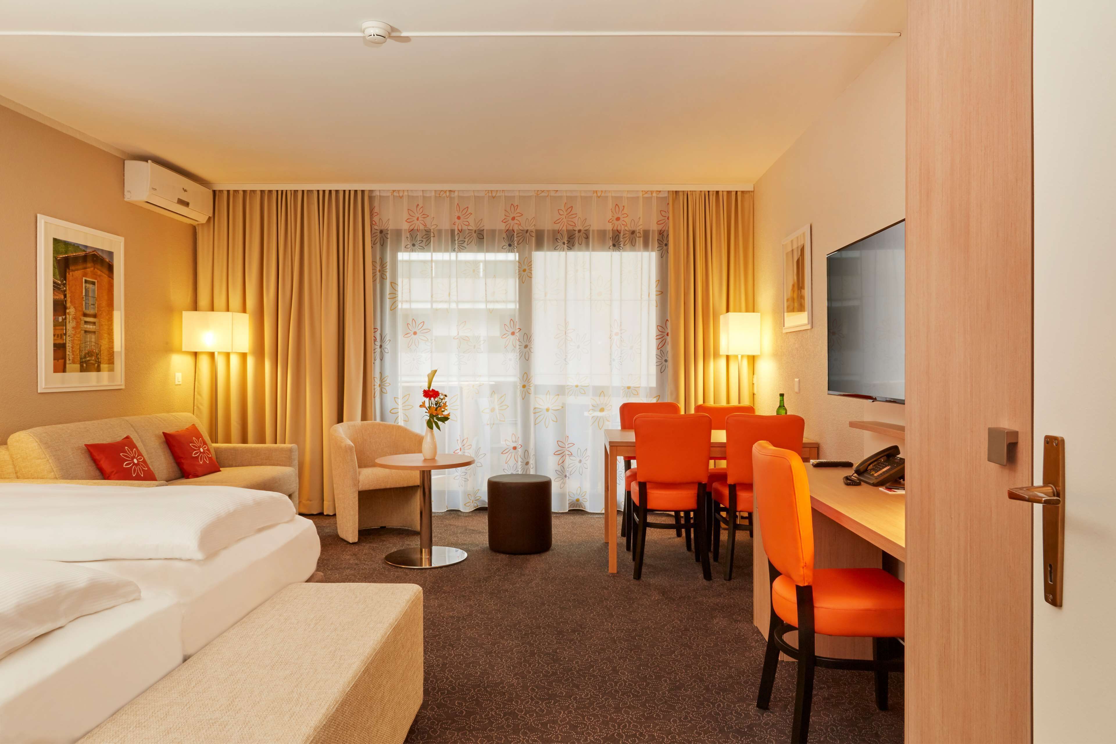 A hotel room with a bed, a desk, two chairs, a small table, a lamp, a TV, and a window with curtains.