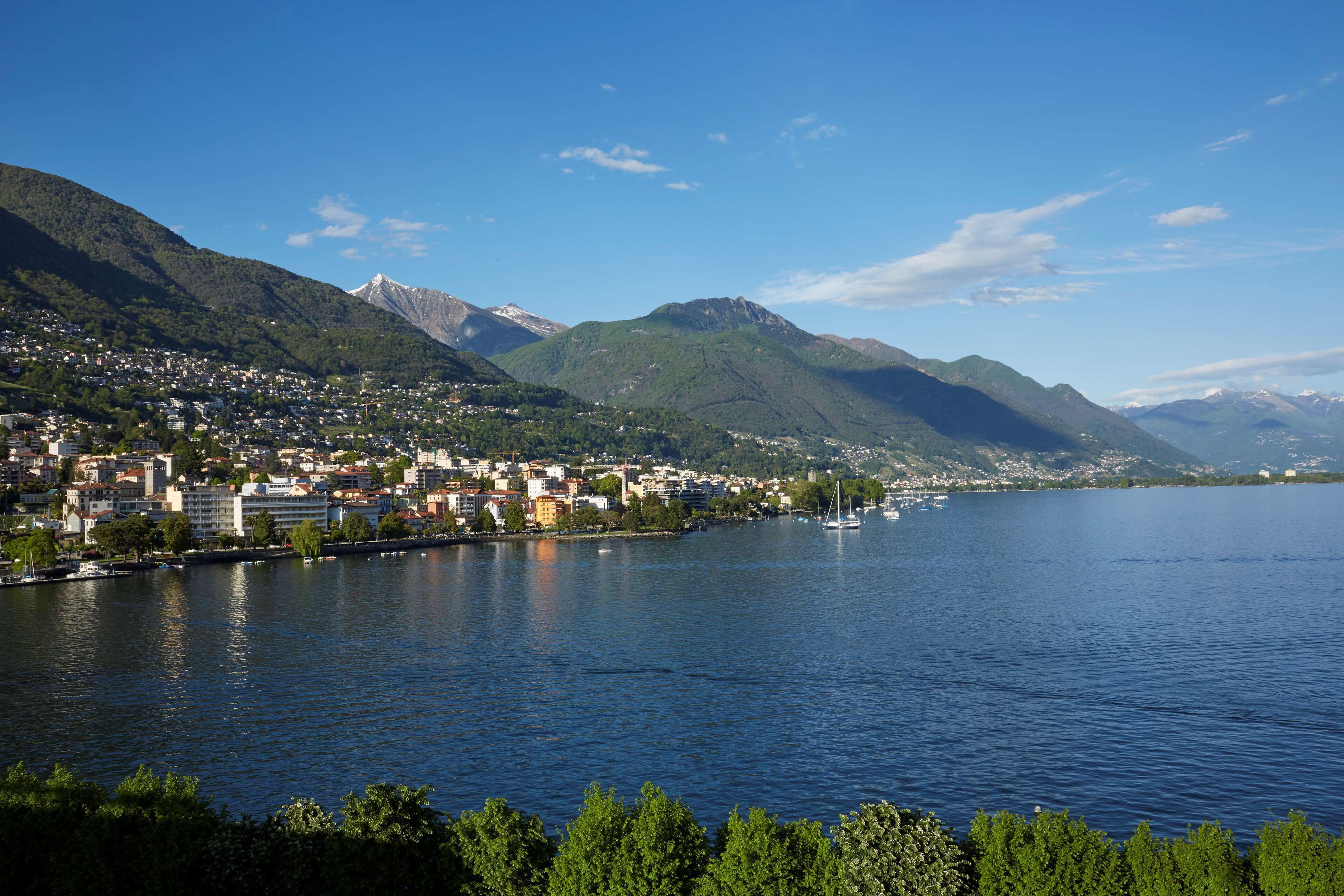 Une ville au bord d’un lac, avec des bâtiments qui bordent la rive, un voilier et des montagnes en arrière-plan.