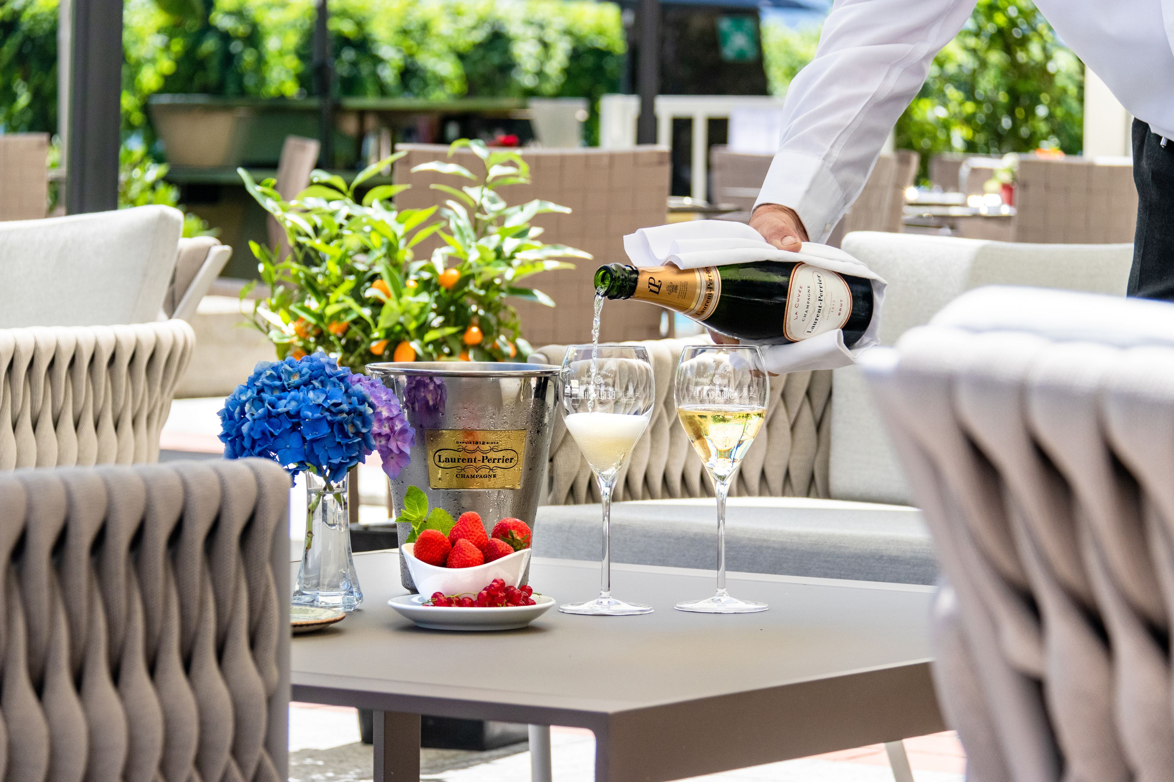 A person pouring champagne into a glass on an outdoor table with a bowl of strawberries and a vase of flowers.