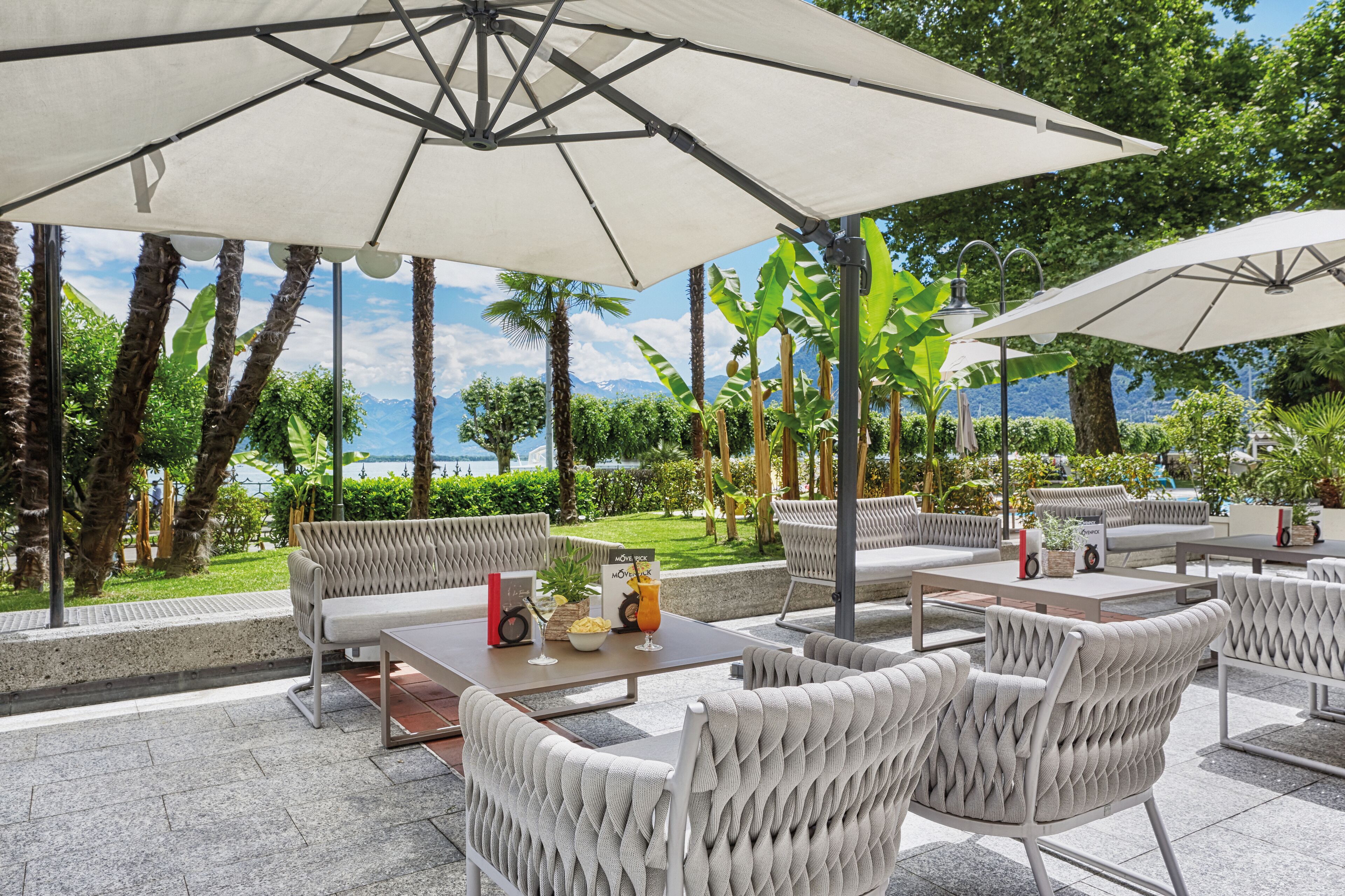 An outdoor seating area with white wicker furniture, large umbrellas, and a view of the sea and mountains.