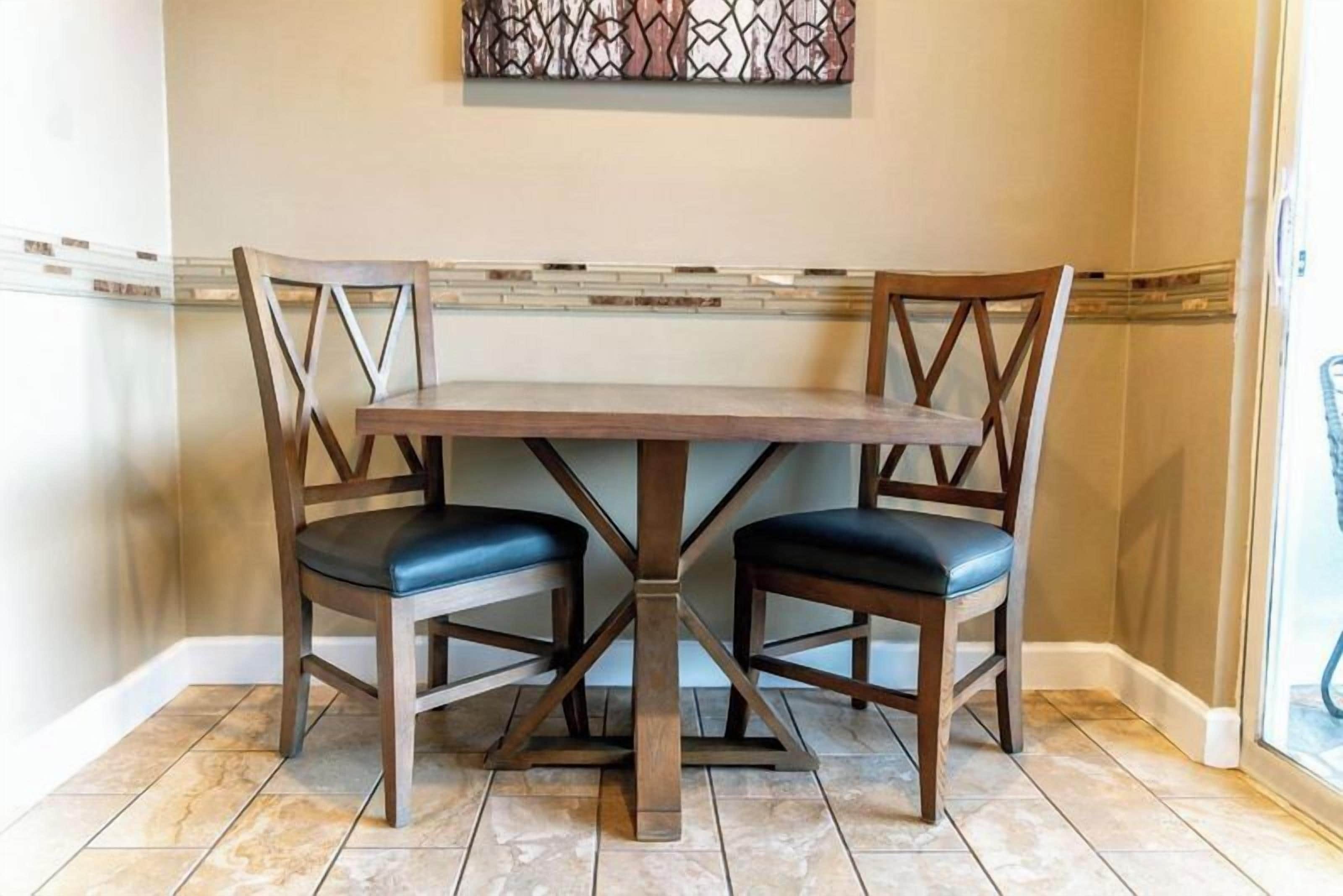 A dining area with a wooden table and two chairs, a decorative wall piece above, and a tiled floor.