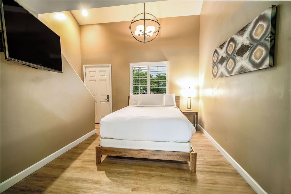 A bedroom with a bed, a TV mounted on the wall, a window with blinds, and a chandelier.