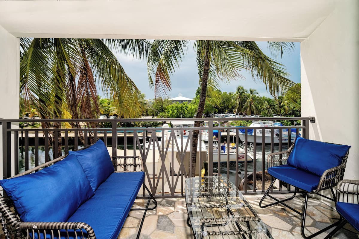 A balcony with wicker furniture and a glass table, overlooking a marina with boats and palm trees.
