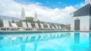 Una piscina con sillas de descanso y sombrillas, un área cercada y un edificio con una puerta.