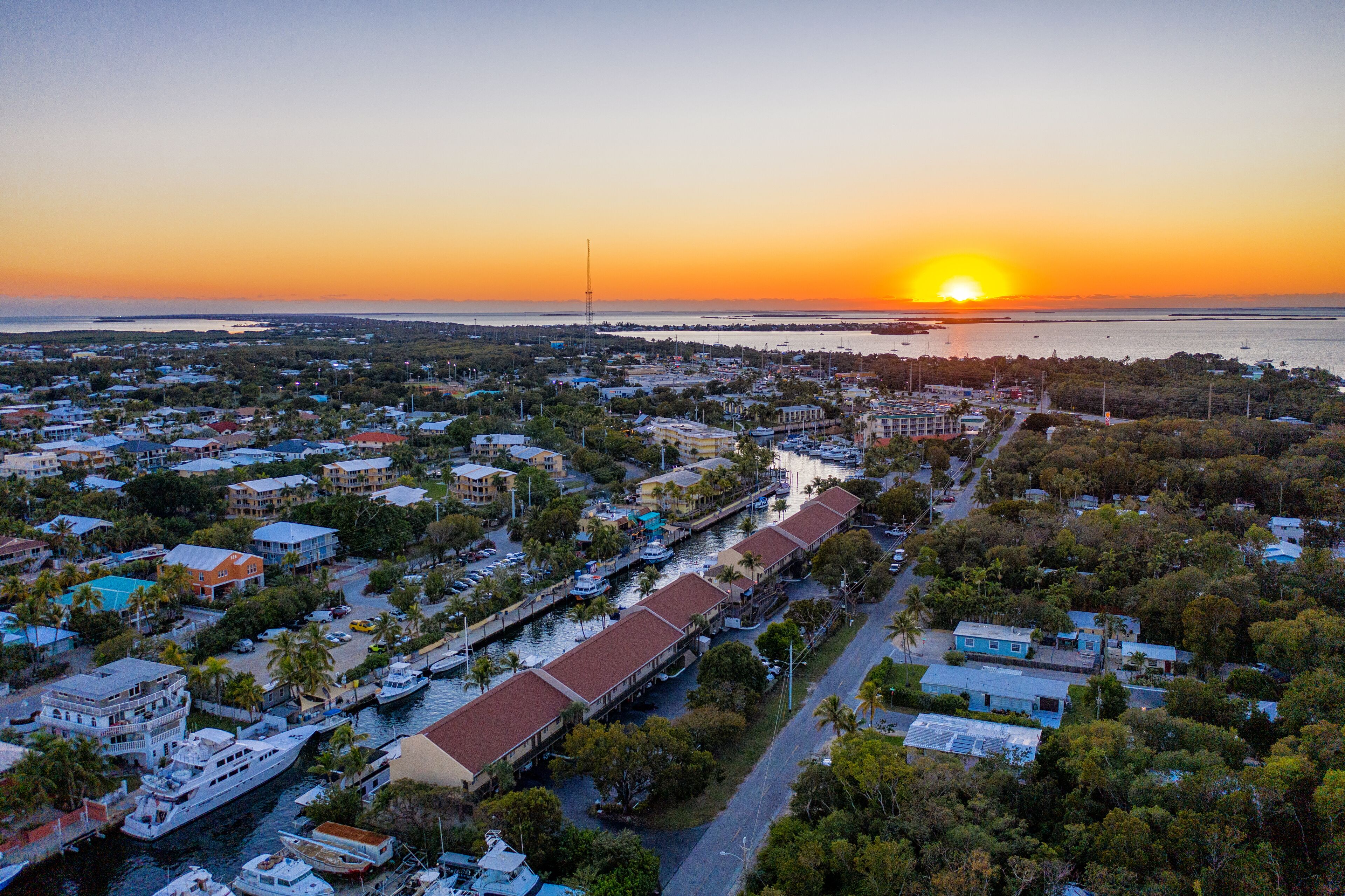 An aerial view of a coastal town with a marina, buildings, and a sunset over the water.