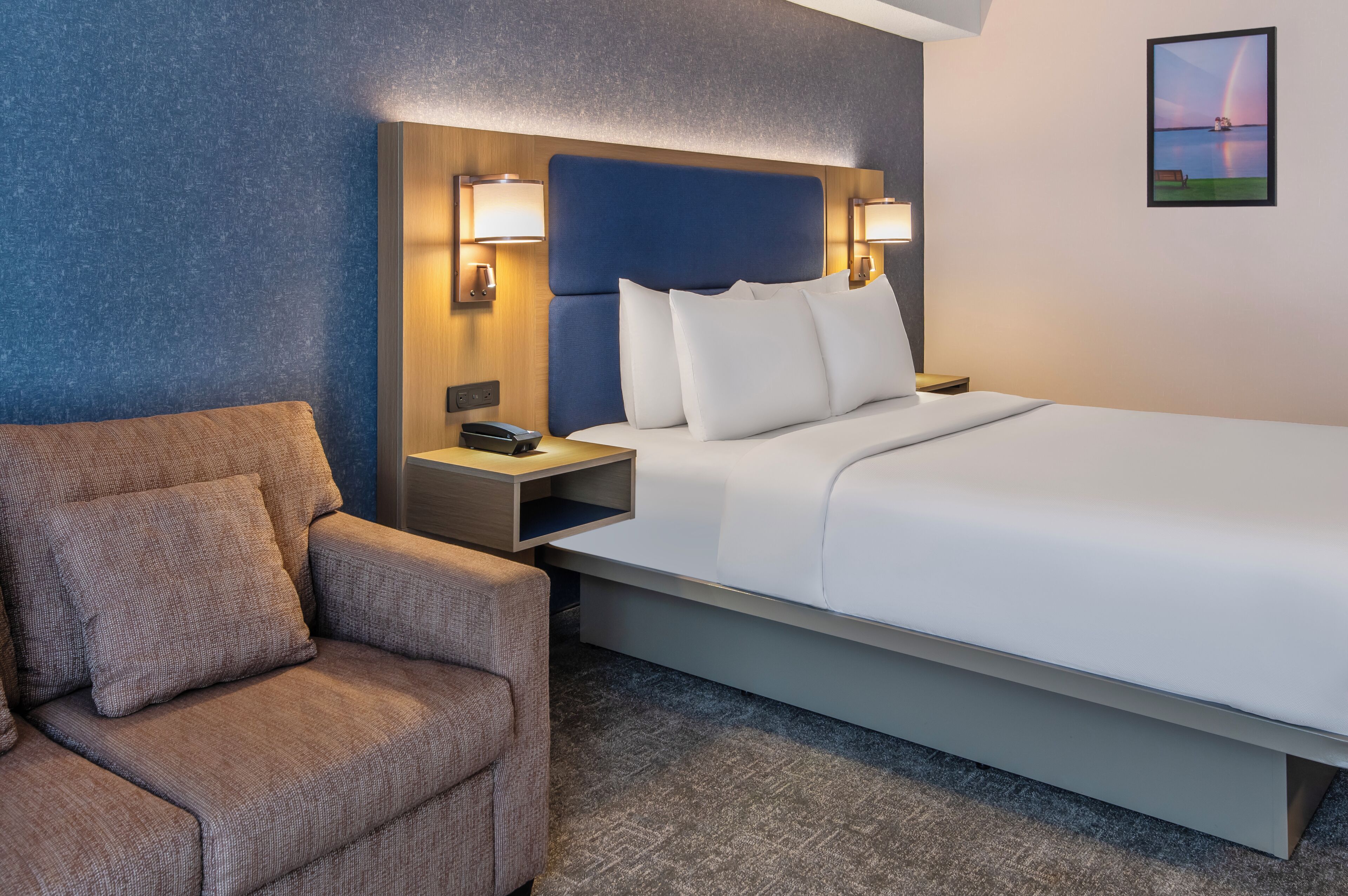 Quarto de hotel com cama, cadeira, mesa de cabeceira e luminária na parede.
