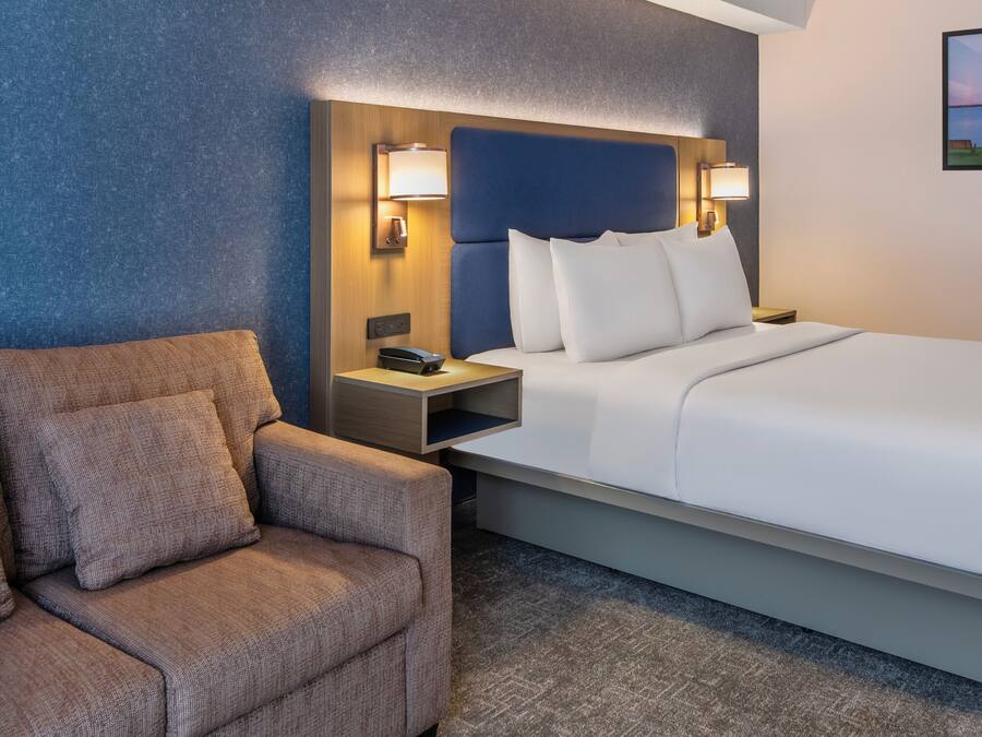 Quarto de hotel com cama, cadeira, mesa de cabeceira e luminária na parede.