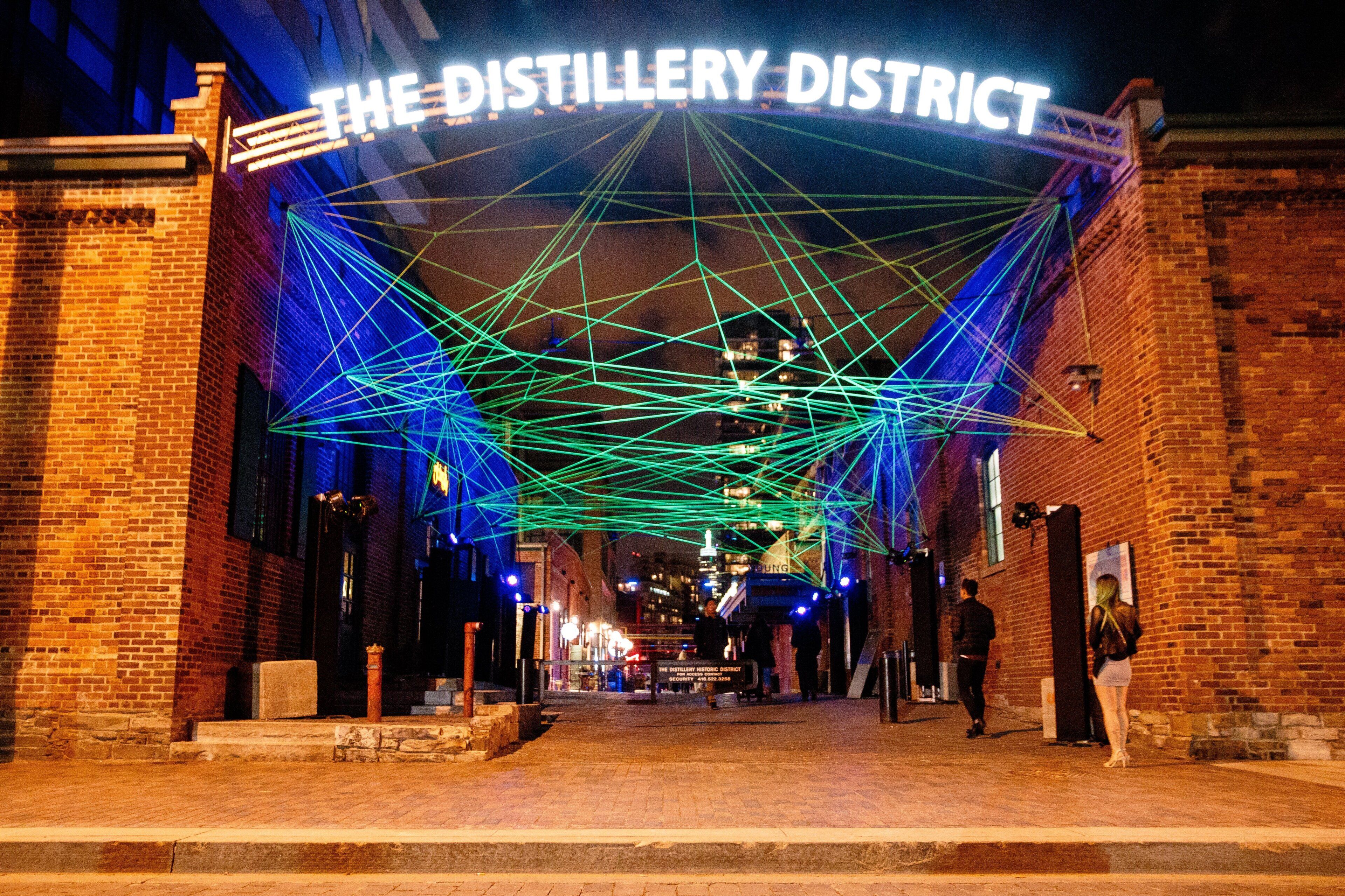 A brick building with a sign reading 'THE DISTILLERY DISTRICT' and a geometric light installation above.
