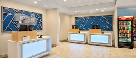 A hotel reception area with two reception desks, a Coca-Cola refrigerator, and a wall with geometric patterns.