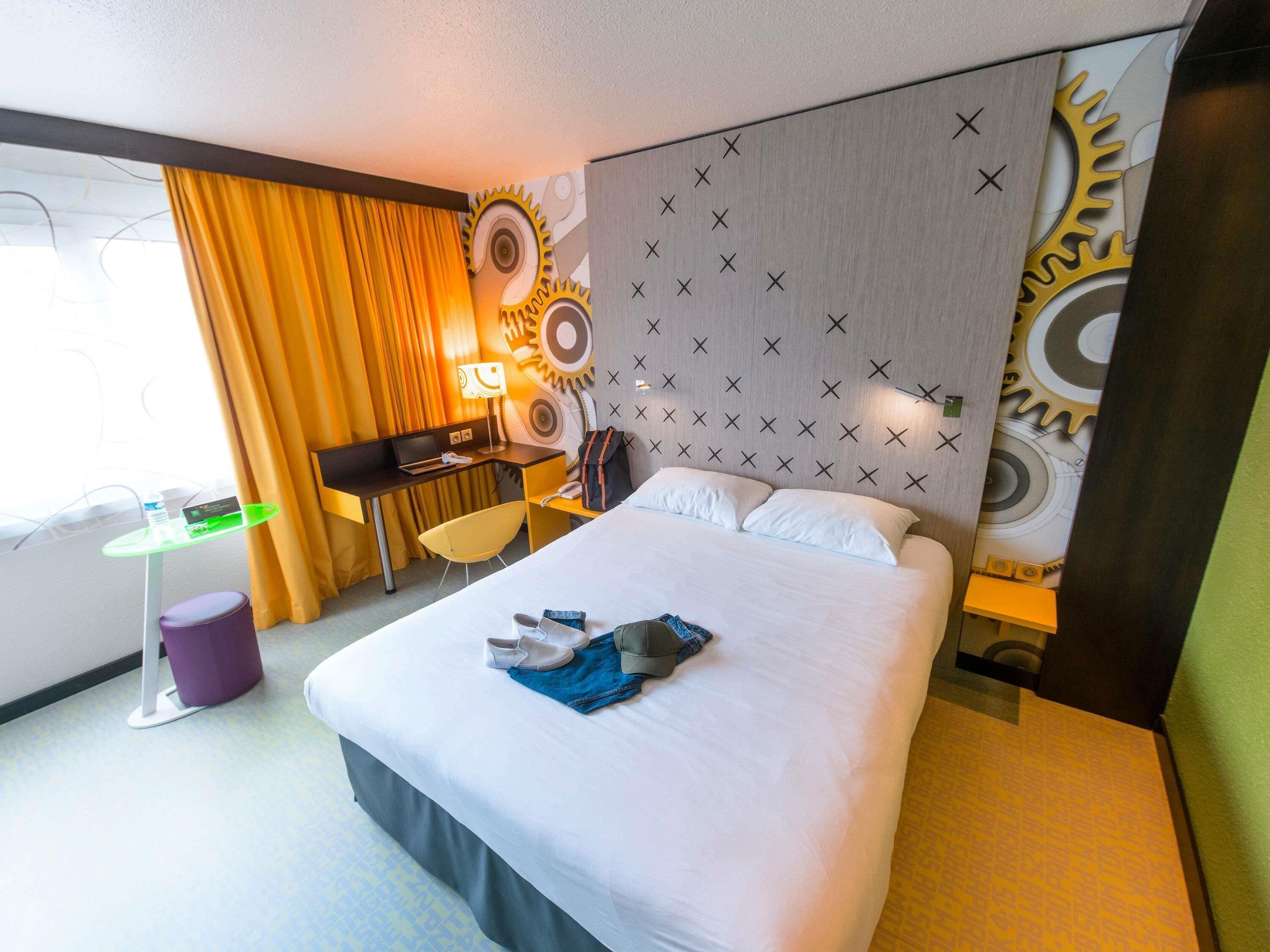 A hotel room with a bed, a desk, a chair, a small table, and a wall with a mechanical design.